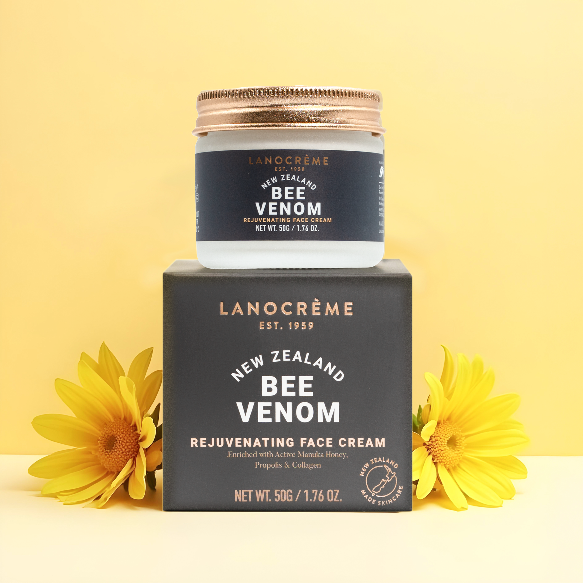 A jar of Lanocrème Bee Venom Rejuvenating Face Cream sits on top of its dark gray box, with two yellow flowers on either side, against a pale yellow background.